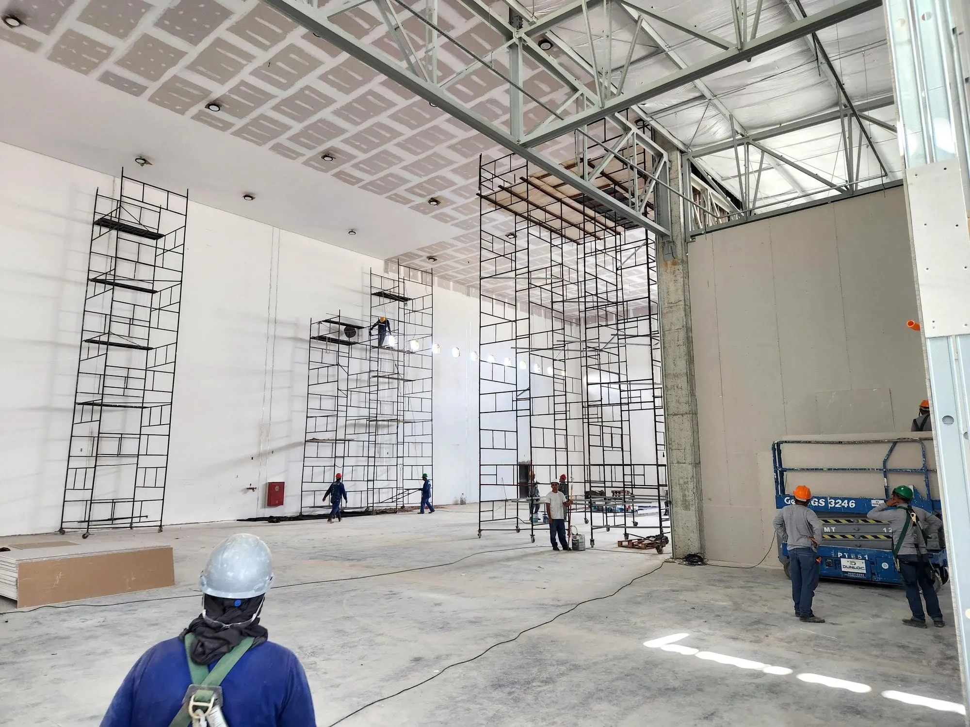 Interior de galpão em obra, com andaimes e equipe de capacete